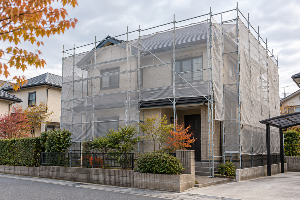 曇り空の下、足場と飛散防止シートに囲まれた住宅のイメージ(AI生成)