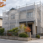 曇り空の下、足場と飛散防止シートに囲まれた住宅のイメージ（AI生成）